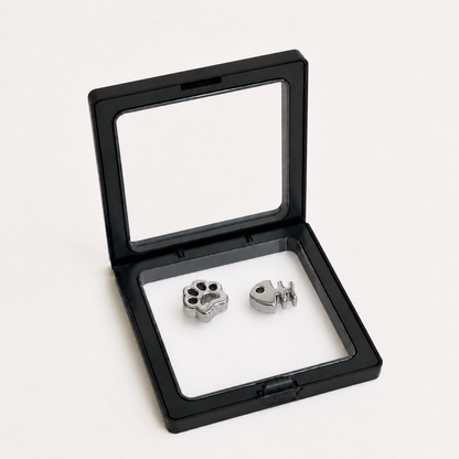 Two silver stainless steel matcha sifter weights, one shaped like a paw print and the other like a fish, displayed inside an open black presentation box with a clear lid on a white background.