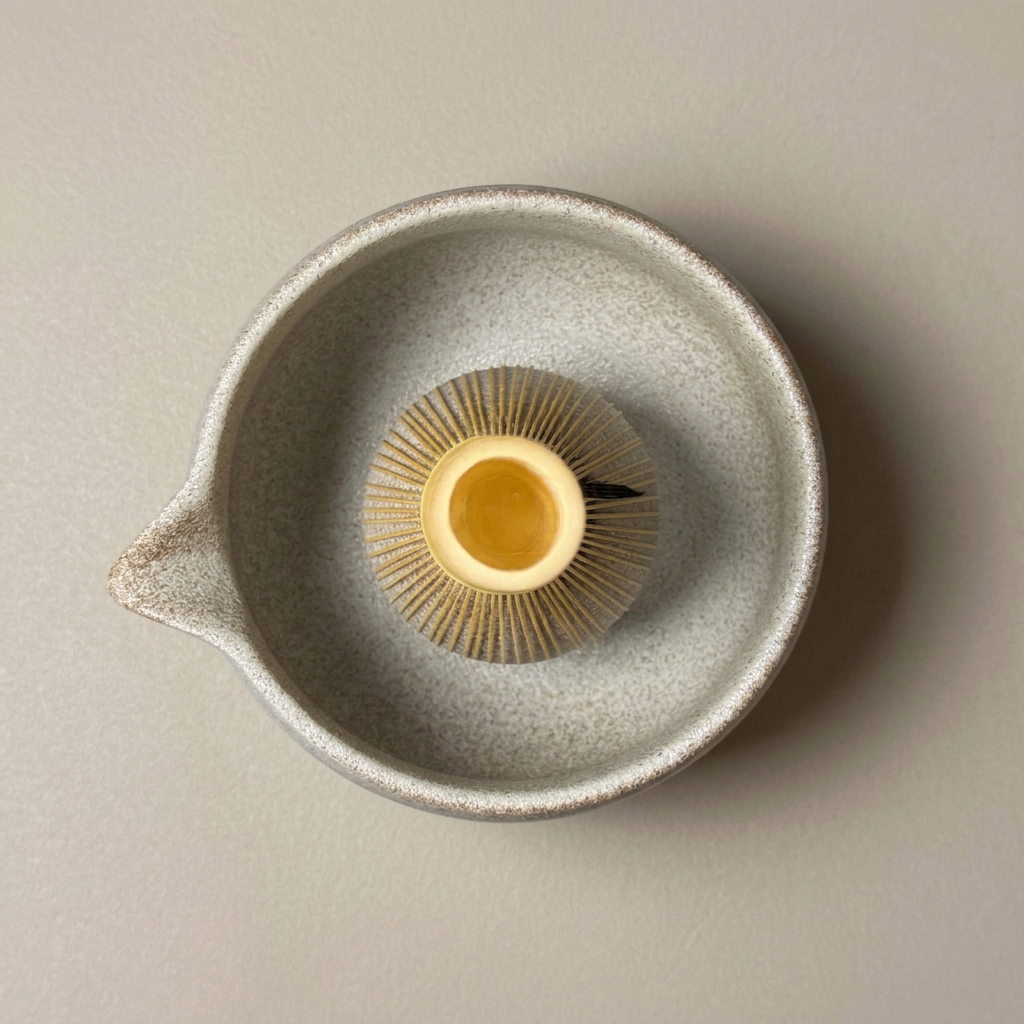 Top view of a golden bamboo matcha whisk with its hollow handle facing upward inside a speckled ceramic bowl.