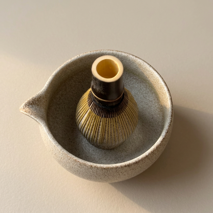 Purple bamboo matcha whisk resting inside a speckled ceramic bowl with a pouring spout.